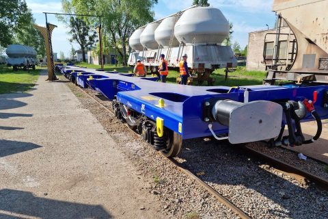 Three quarter trackside view of a new flatbed intermodal carrier wagon going through the final gauge clearance hoop on a sunny day with engineers and trees in background