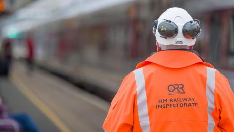 Inspector in freight yard with back to camera wearing orange Office of Rail and Road overcoat and helmet