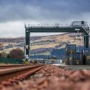 A track level view of the overhead crane at Highland Spring Bottling Plant terminal in Scotland. The red ballast contrasts with the heather on the hills and the grey skies overhead